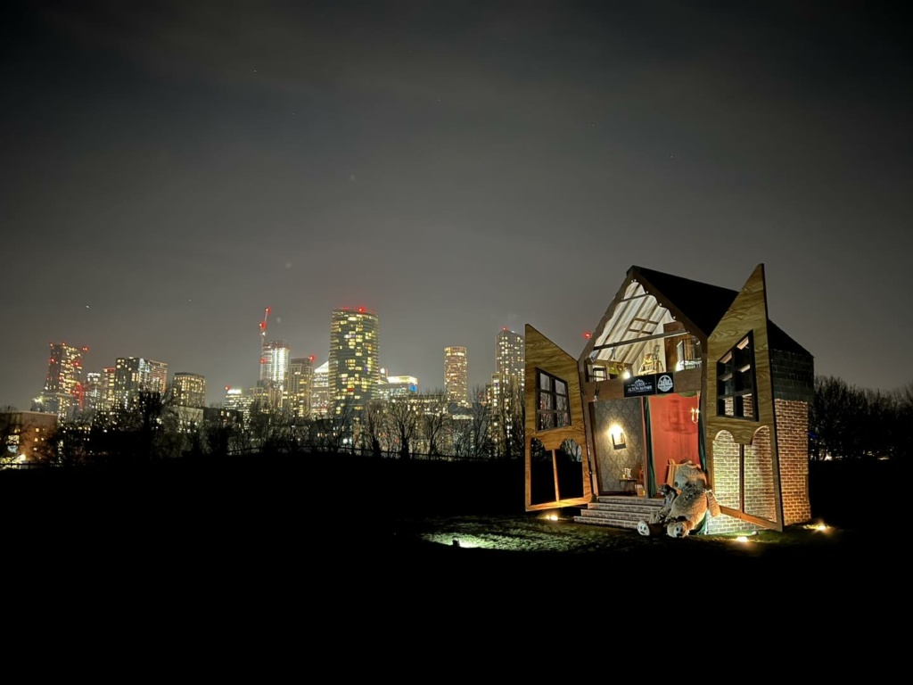 A larger than life dolls house sits in a field with the skyline of Canary Wharf in the background.